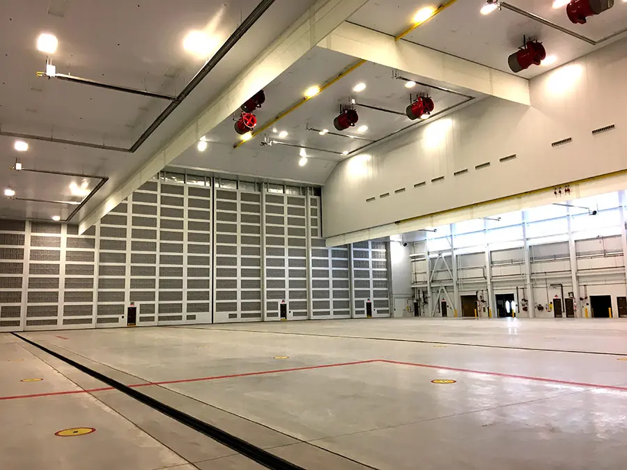 Spacious, well-lit industrial hangar with high ceilings, red overhead cranes, large closed sectional doors, and a vast, clean concrete floor.