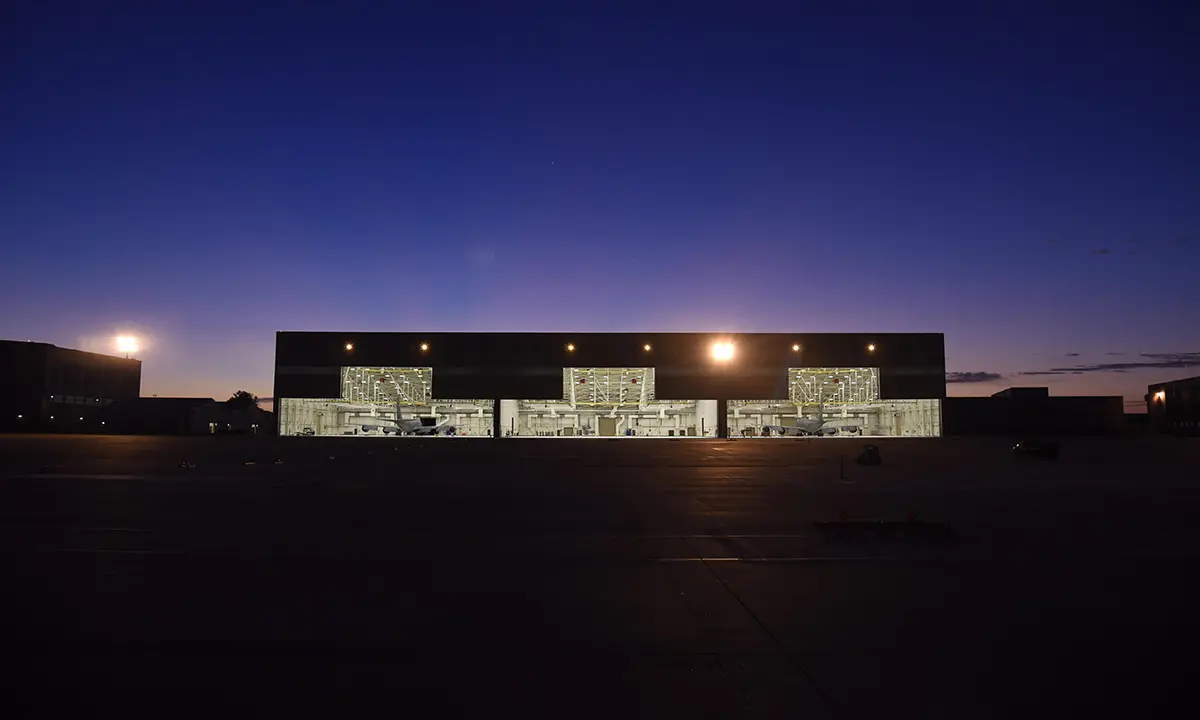 A large aircraft hangar glows brightly at night under a deep blue sky. The hangar's open doors reveal illuminated interiors, creating a stark contrast.