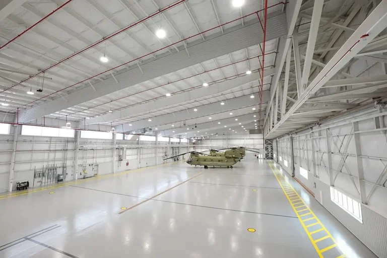 Spacious hangar interior with high ceilings, bright lights, and a yellow-striped floor. A military helicopter sits at the center, conveying readiness and precision.