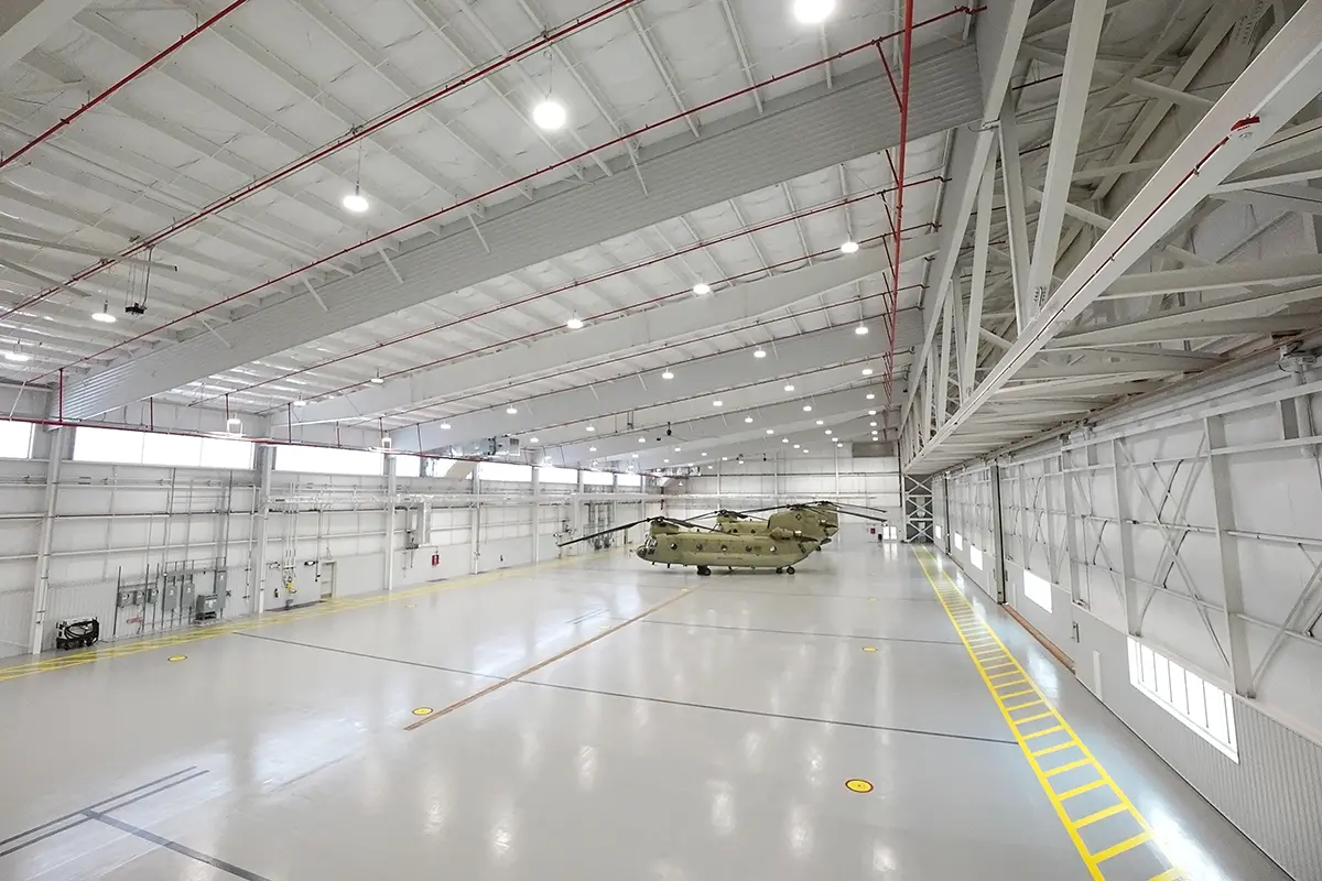 Spacious hangar interior with high ceilings, bright lights, and a yellow-striped floor. A military helicopter sits at the center, conveying readiness and precision.