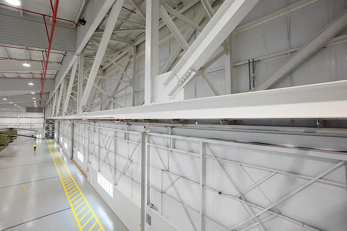 Wide-angle shot of a large, empty aircraft hangar with high ceilings and exposed steel beams. Yellow lines on the floor guide a distant worker.