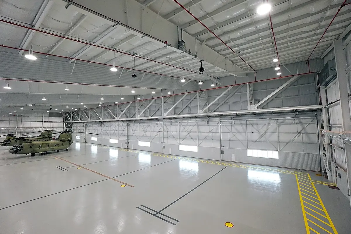 Spacious hangar interior with a gray floor marked with yellow lines and lights on the ceiling. Two helicopters are parked on the left.