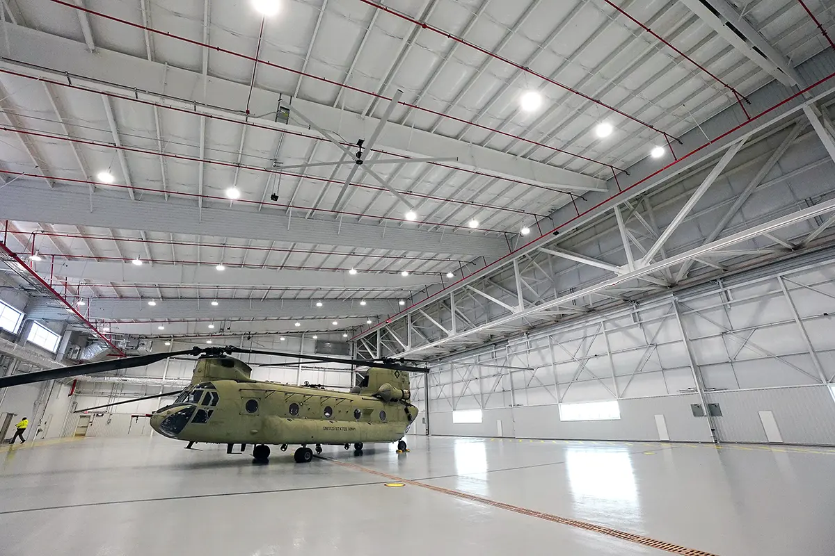 A large, olive-green military helicopter is centered inside a spacious, well-lit hangar with high ceilings and metal beams, creating an orderly and calm atmosphere.