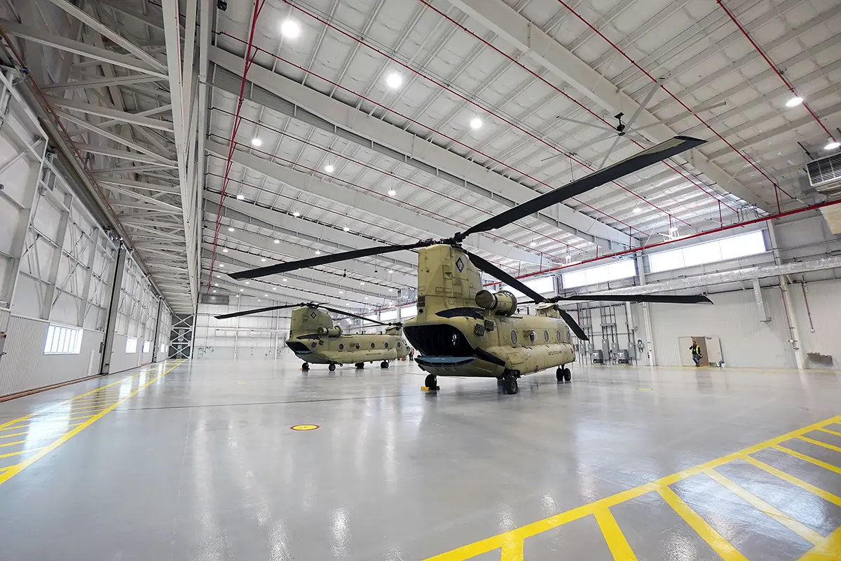 Two tan military helicopters are parked inside a spacious, well-lit hangar with a high ceiling. The floor is smooth with yellow safety markings.