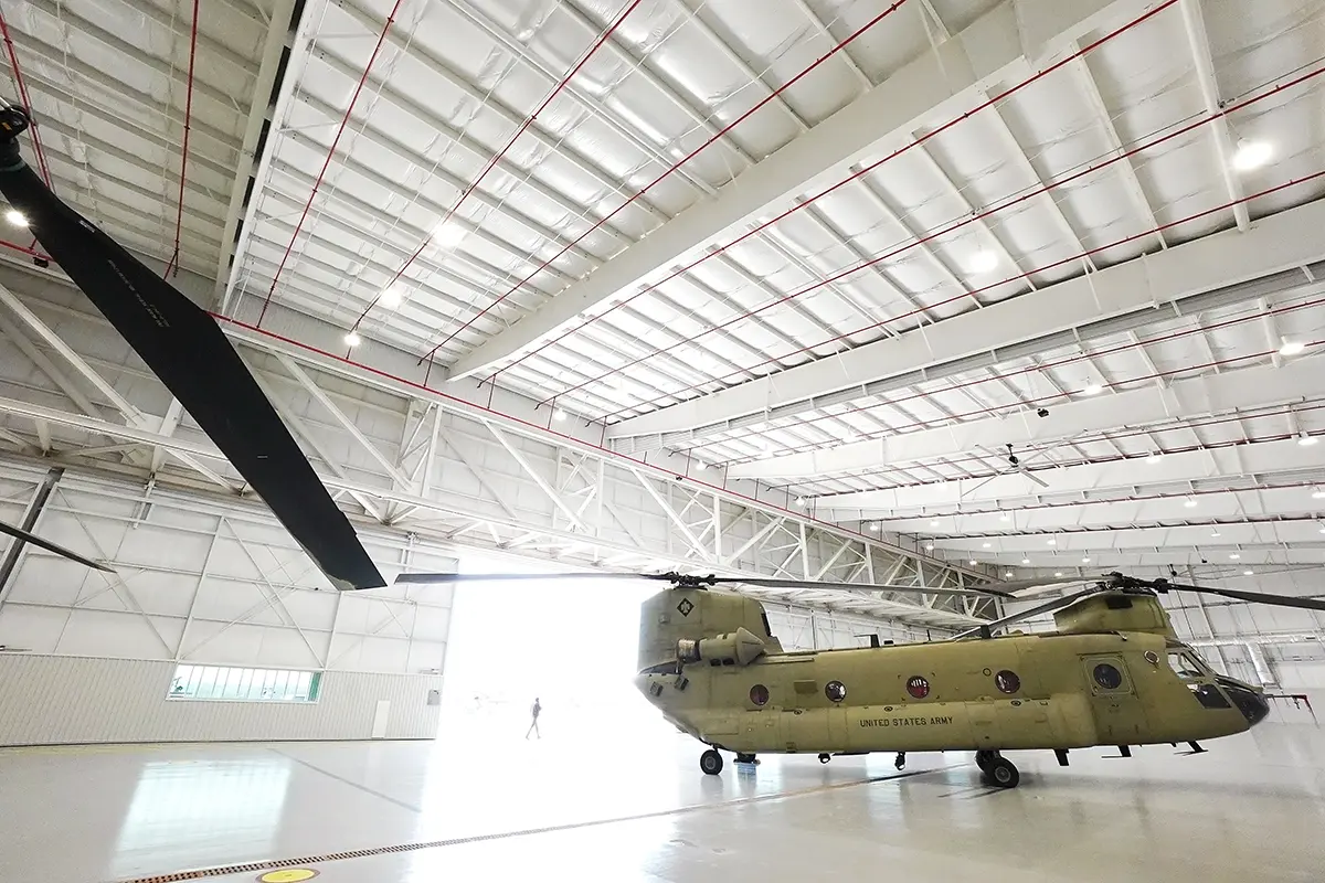 A military helicopter is parked in a large, bright hangar with a high ceiling. Its rotors are visible; a person walks near the open hangar door.
