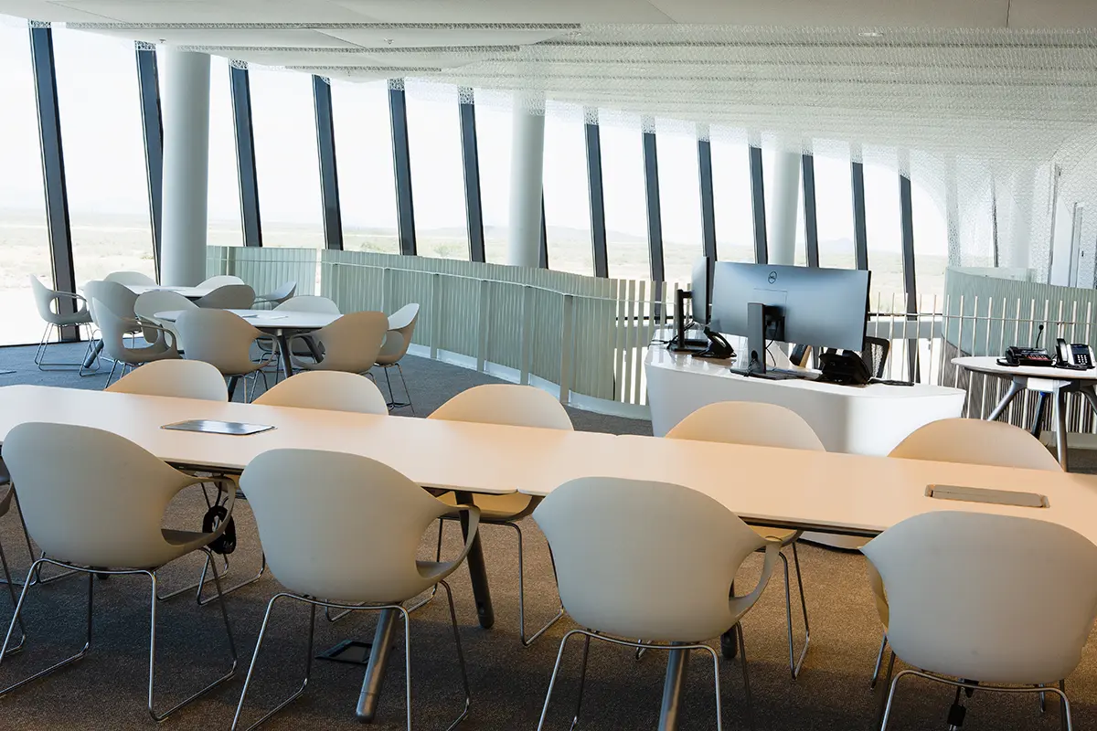 Modern office with large windows, white tables, and chairs arranged in a spacious, open layout. A computer station is visible, creating a calm, professional atmosphere.