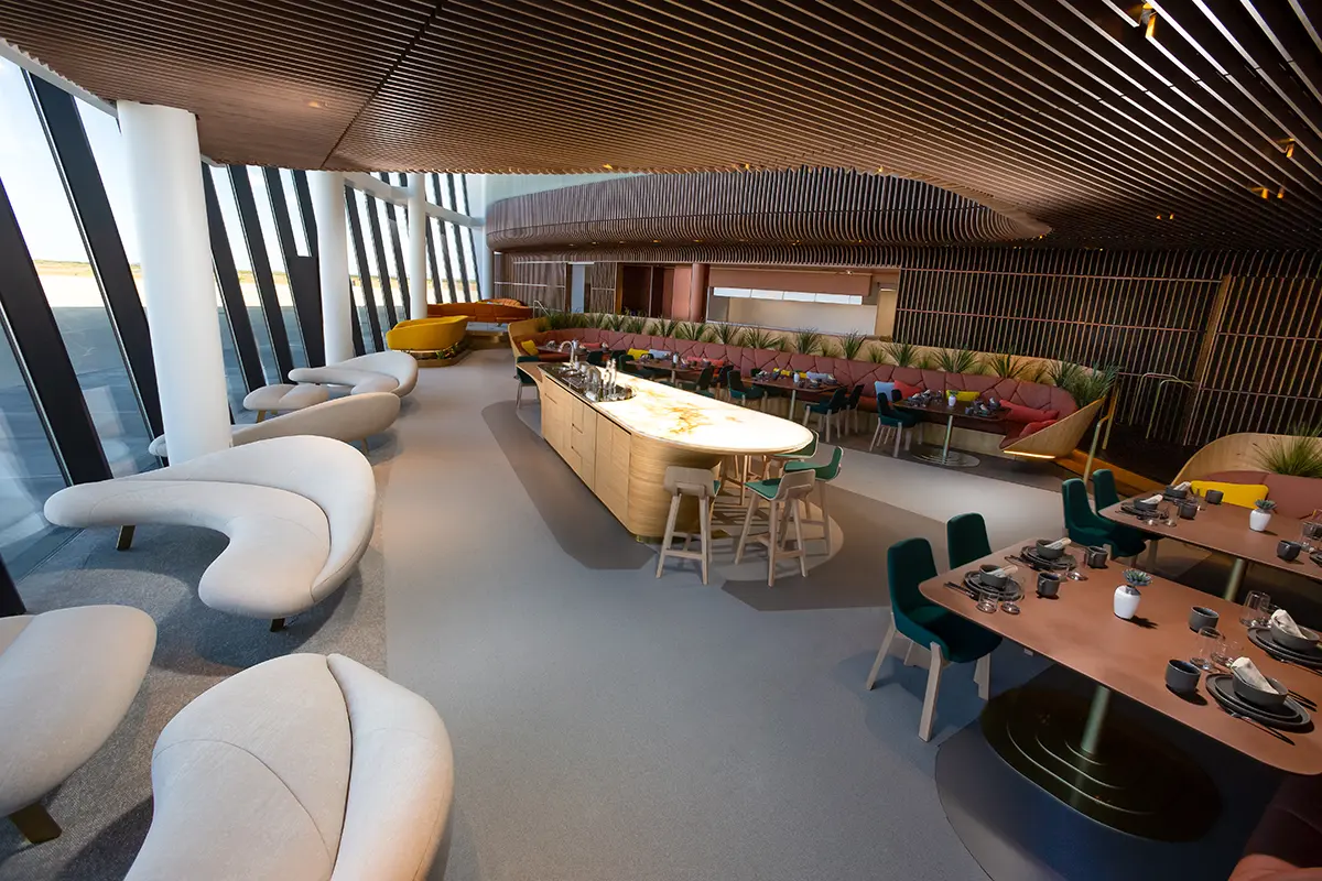 Modern lounge with curved beige sofas, a central bar with light wood and a glowing countertop, and dining tables set with dishes, under a wooden slatted ceiling.