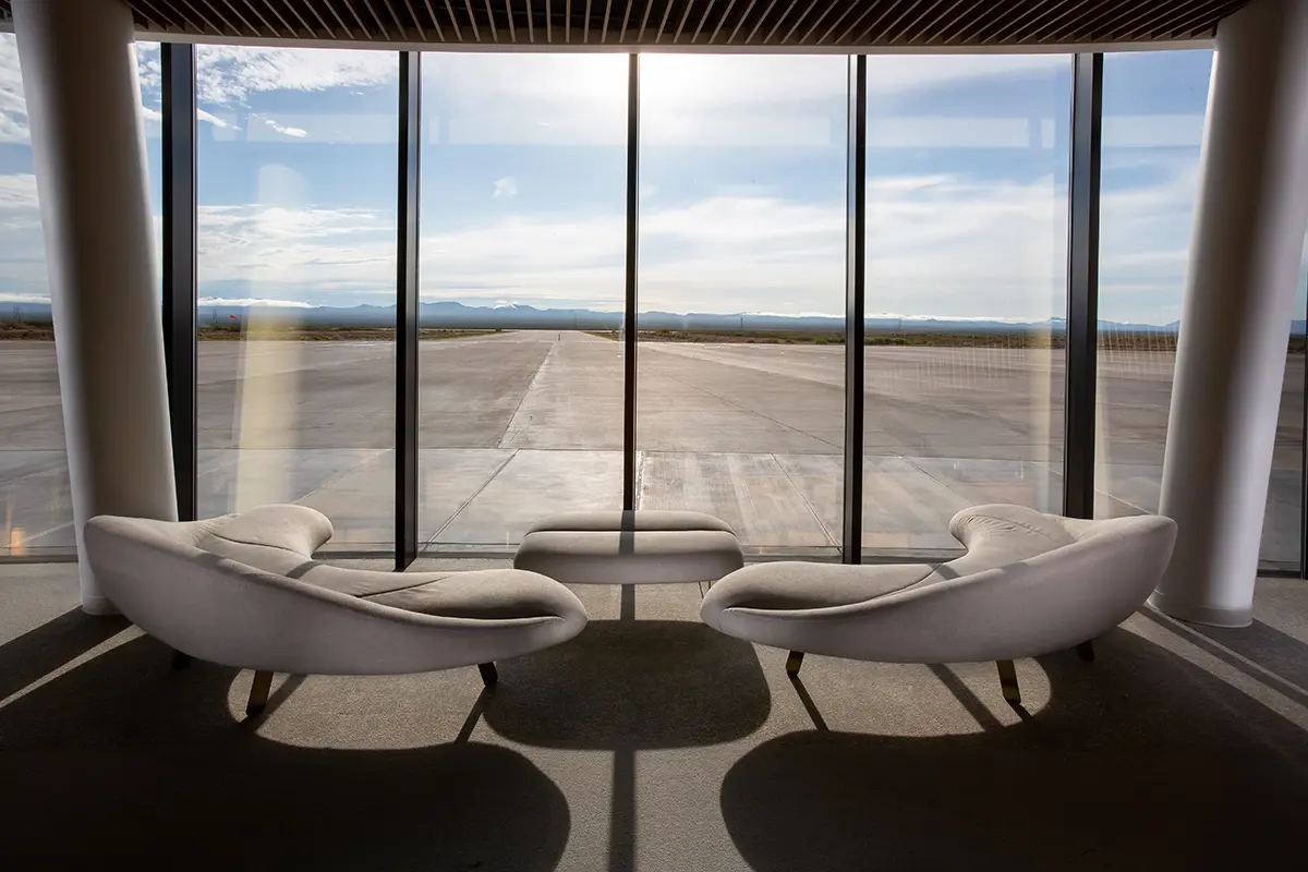 Modern lounge area with two curved, white sofas and a small table by large floor-to-ceiling windows, overlooking a sunny, empty tarmac and distant mountains.