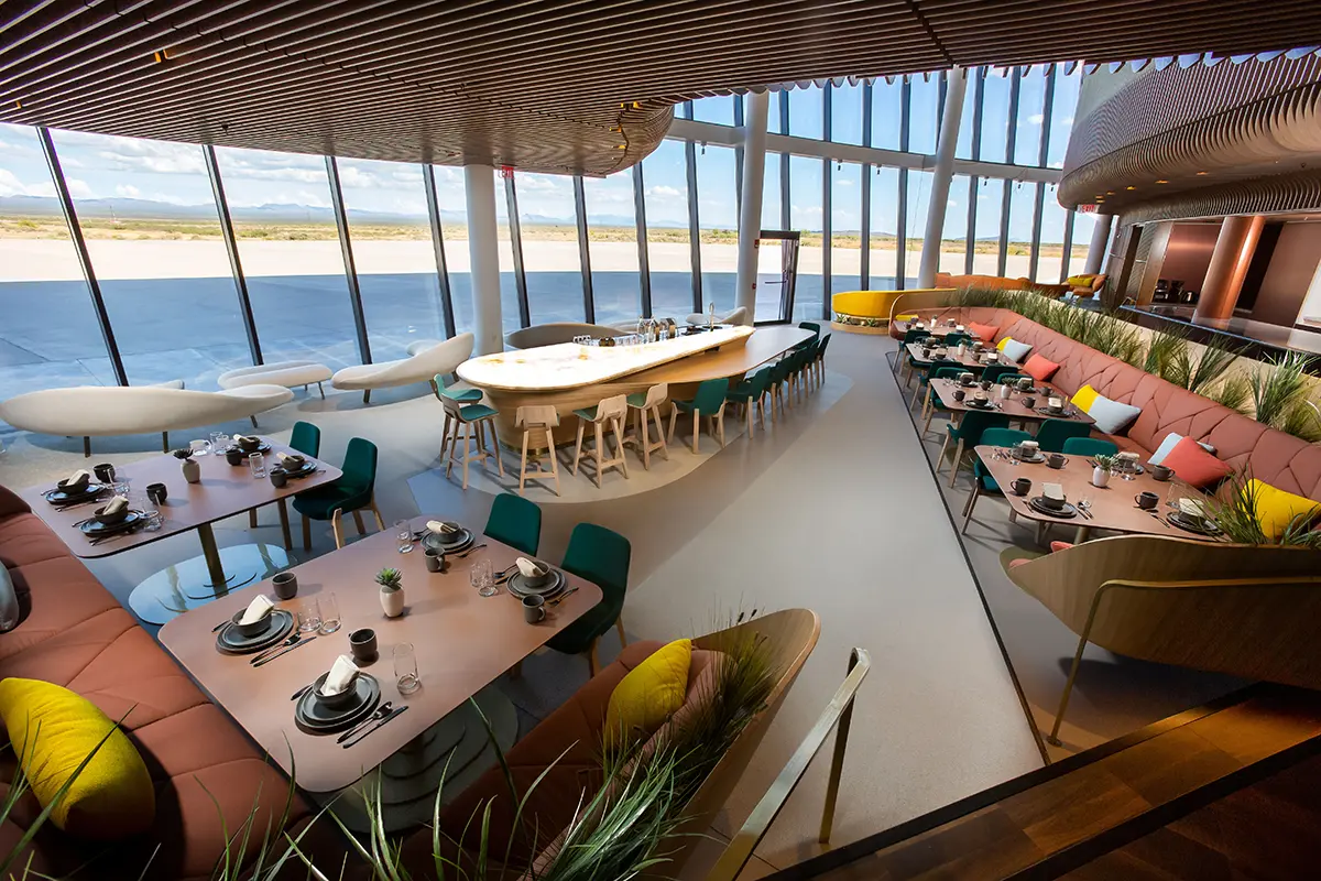 Modern airport lounge with panoramic windows and sleek furniture. Tables are set with dishes, and colorful cushions add a lively touch. Bright, airy ambiance.