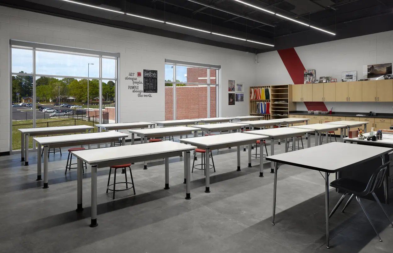 Wide, well-lit classroom with large windows, white tables, and red stools. Art supplies line the shelves, creating a creative and inviting atmosphere.