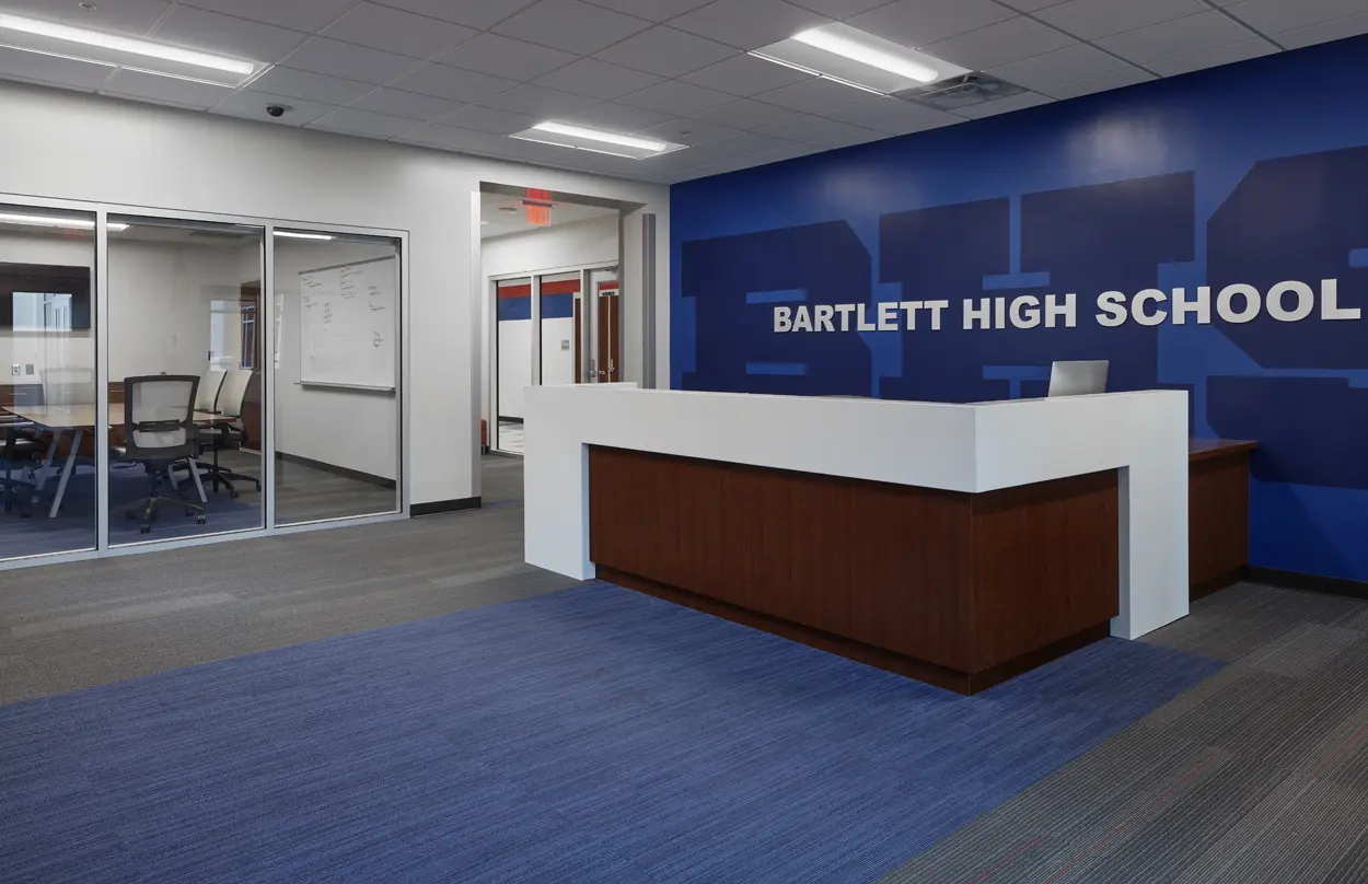 Modern high school lobby with a white reception desk against a blue wall displaying "Bartlett High School." Glass-walled meeting room visible on the left.