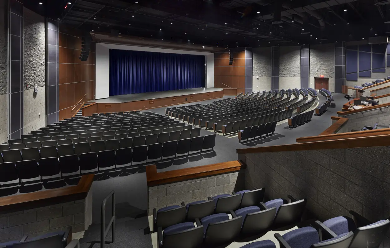 A modern, empty auditorium with blue cushioned seats faces a stage with a closed blue curtain. The room has a calm, anticipatory feel.