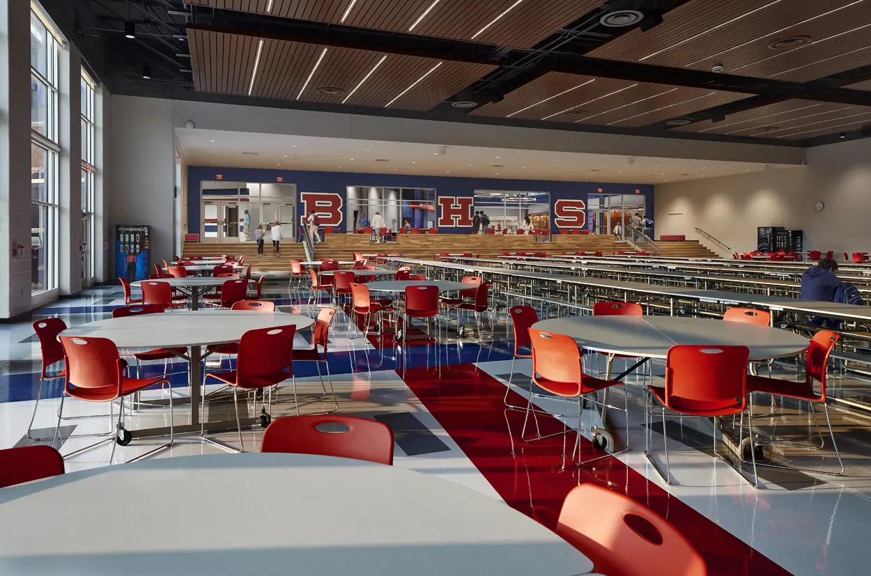 A spacious school cafeteria with red chairs and tables on a polished floor. The letters "BHS" on the back wall, and people walk by, creating a lively, open atmosphere.