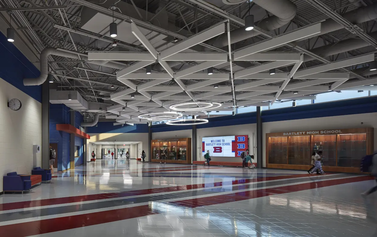 Spacious school lobby with geometric ceiling, red and gray tiled floor. People walk by trophy cases and "Welcome to Bartlett High School" sign.
