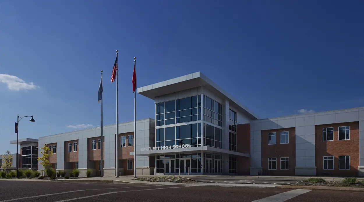 A modern high school building with large windows under a clear blue sky. Three flags fly in front, adding a formal touch. The atmosphere is calm and inviting.