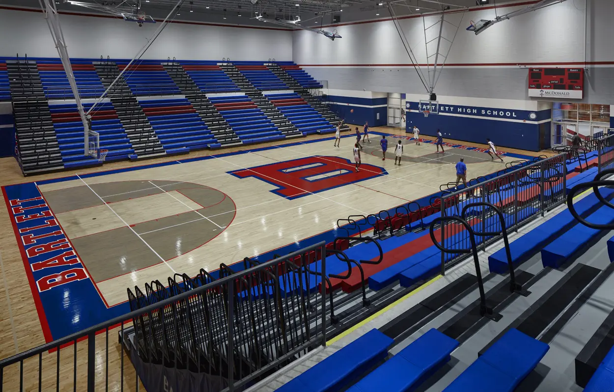 A high school gym with blue bleachers and a large "B" logo on the polished wood floor. Students are playing basketball, creating an energetic atmosphere.