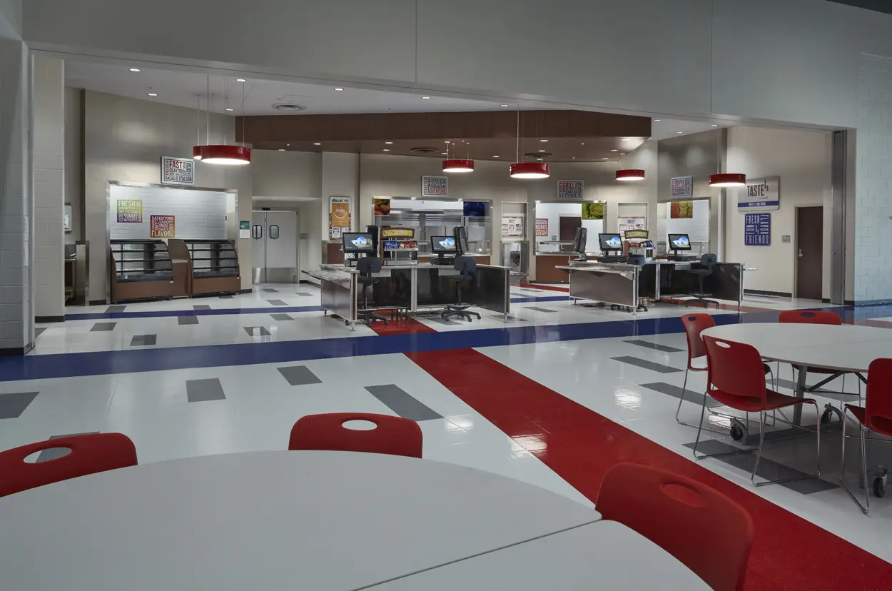 Spacious cafeteria with modern design, featuring red and gray accents. Tables with red chairs in the foreground and a food service area in the back.