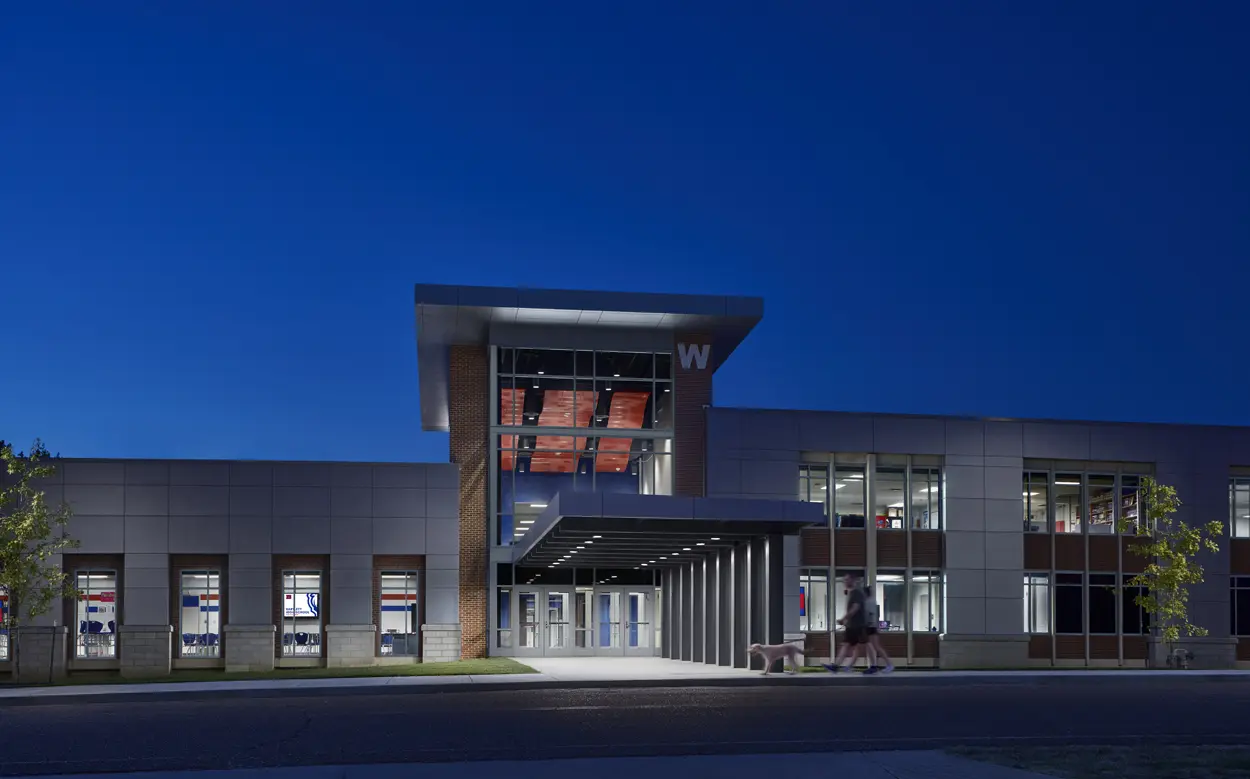 A modern school building at dusk, illuminated by exterior lights. Large windows reveal a colorful interior. A dark blue sky enhances the structure's sleek design.