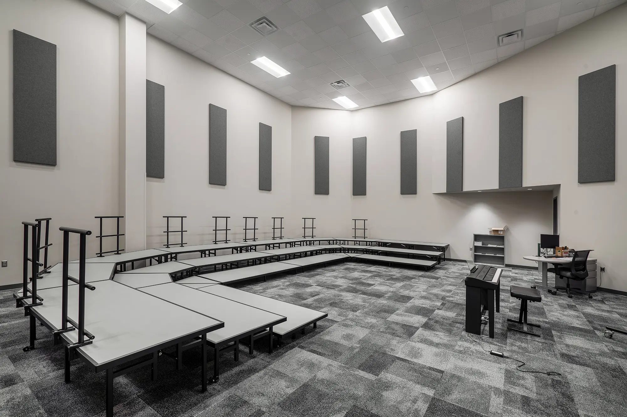 Spacious music rehearsal room with tiered risers, acoustic panels on walls, and a keyboard in the foreground. Neutral tones, organized and quiet ambiance.