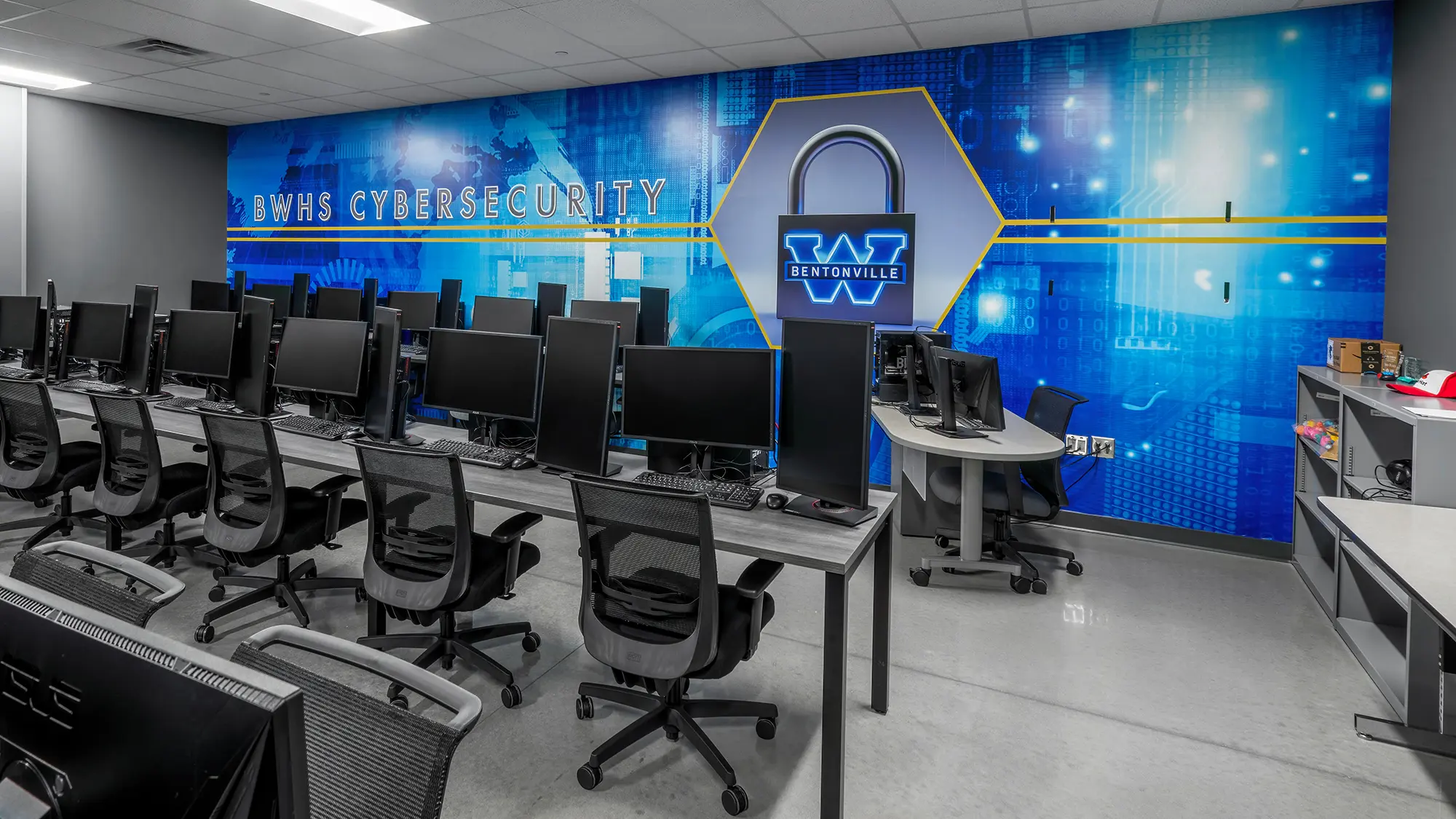 Computer lab for cybersecurity at BWHS, with rows of monitors and chairs. The blue wall displays cyber-themed graphics and a large lock emblem.