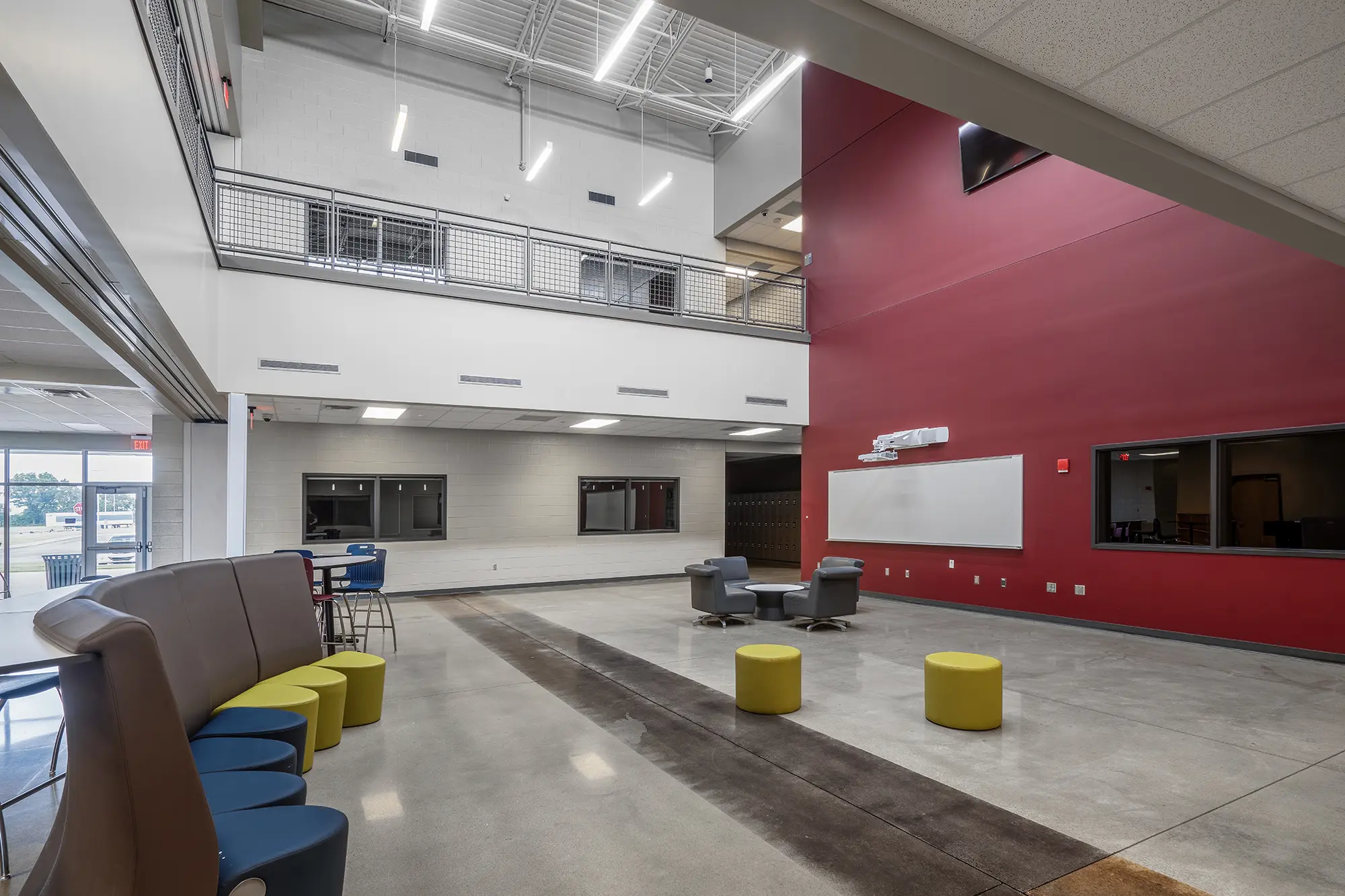 Modern open space room with high ceilings, featuring red and white walls. Includes blue and yellow seating, gray chairs, a whiteboard, and large windows.