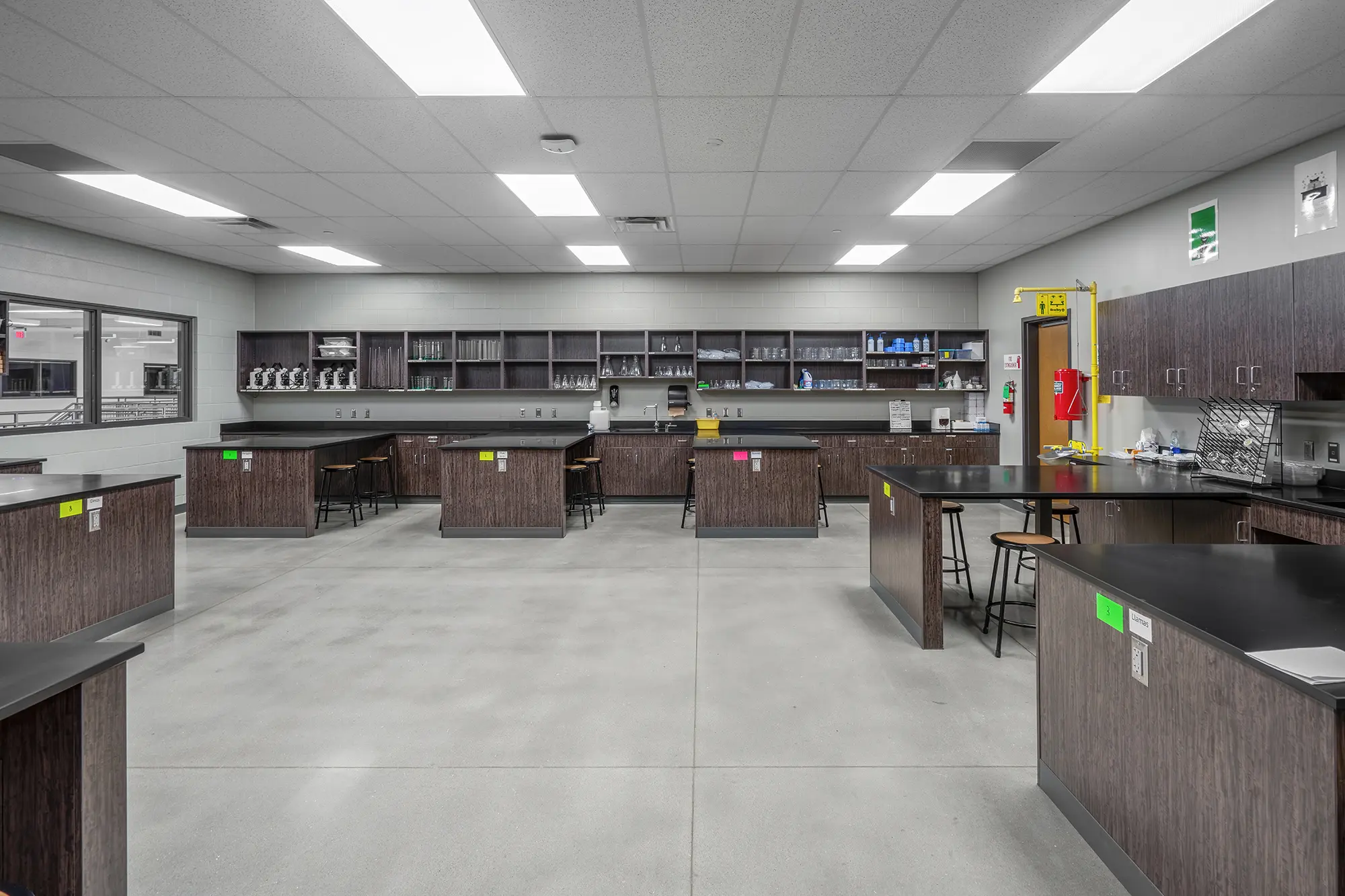 Spacious science lab with dark wooden tables, stools, and labeled cabinets. Bright lighting and organized equipment create a clean, functional atmosphere.
