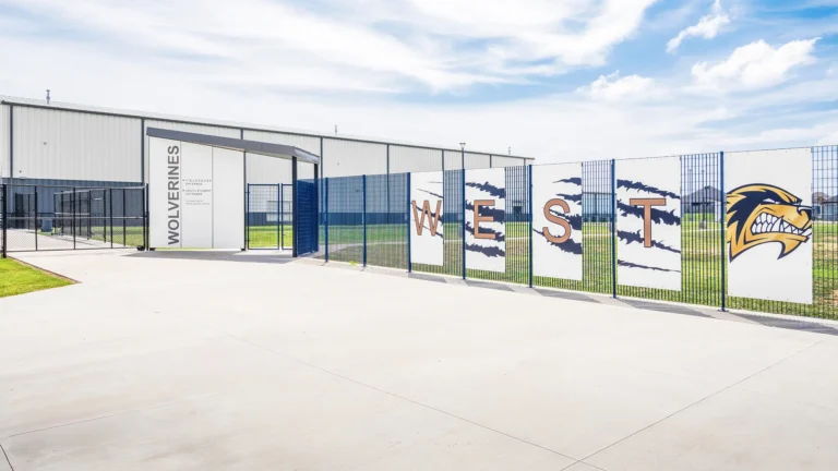 Outdoor view of a sports facility entrance with a sign reading "Wolverines" and metal fencing adorned with art depicting the word "WEST" and a fierce wolverine logo.