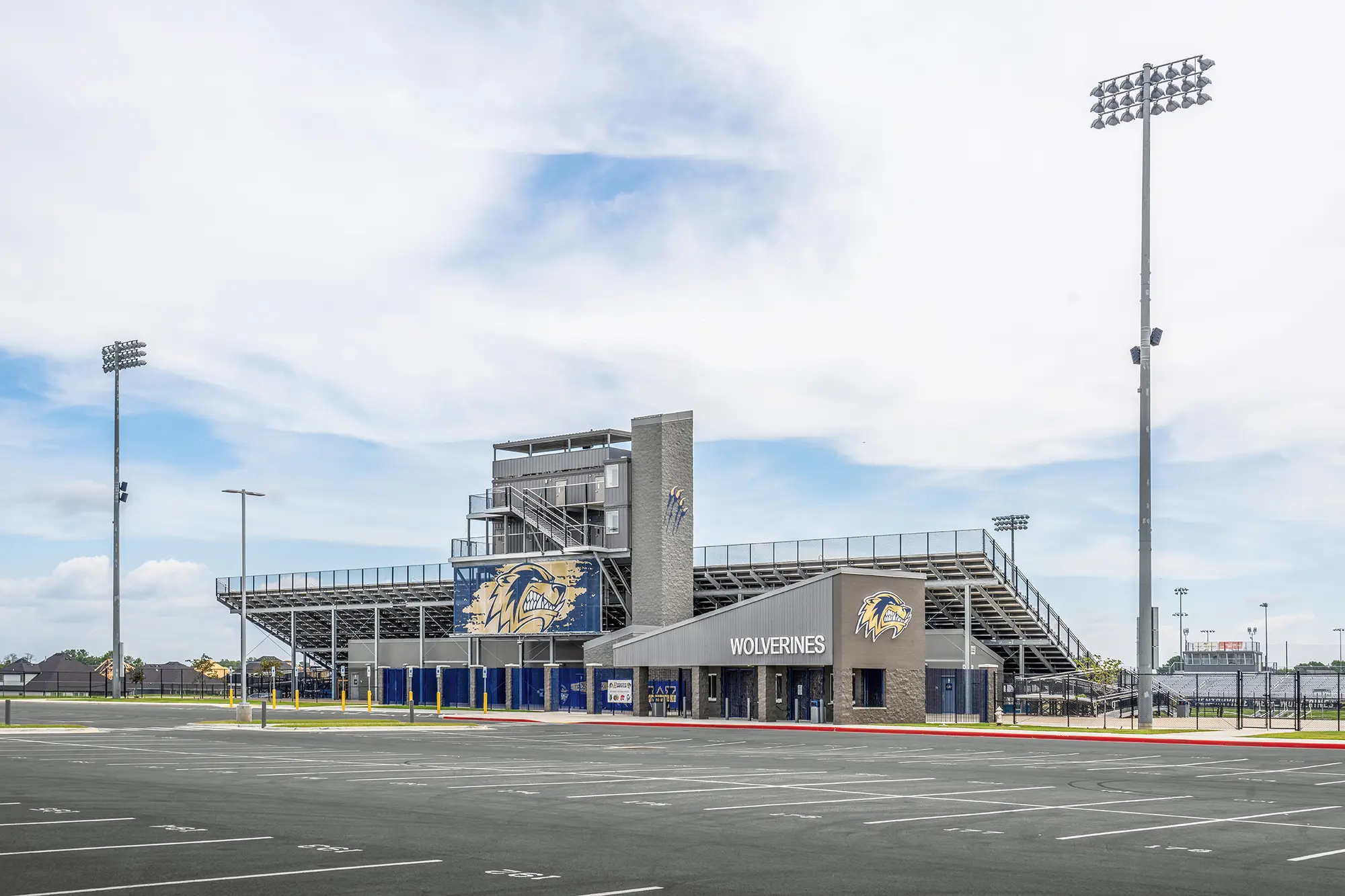 Modern football stadium with empty parking lot, tall floodlights, and an entrance labeled "Wolverines." Sky is bright with scattered clouds, creating a serene atmosphere.