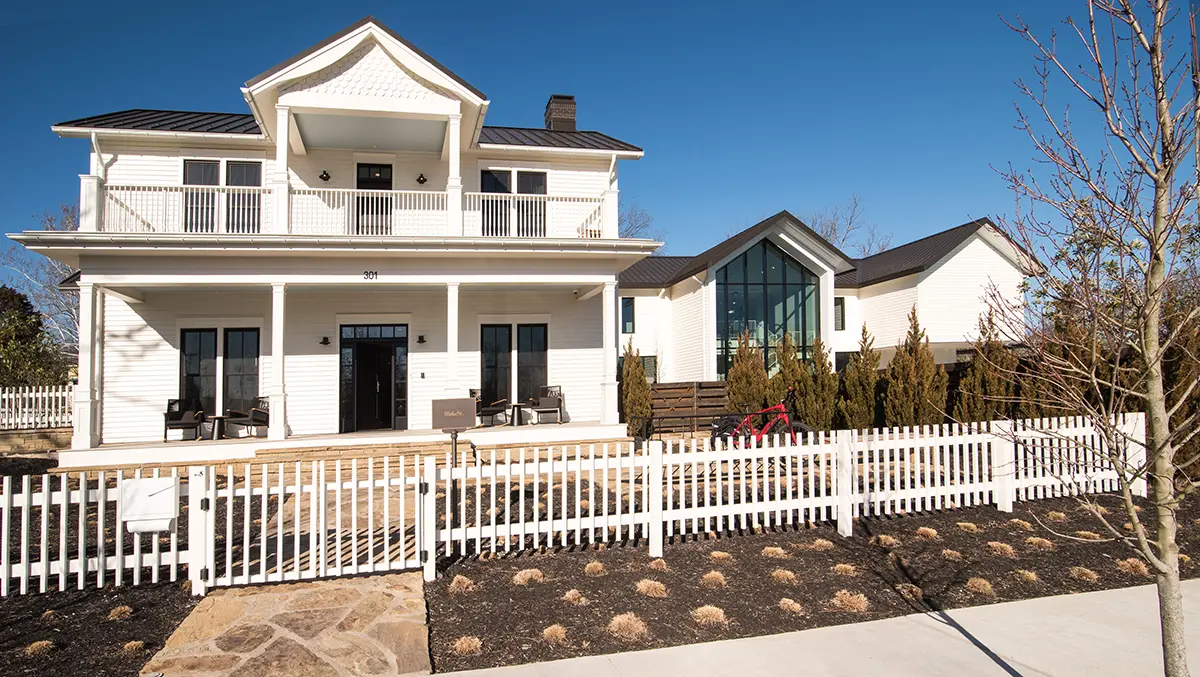 Two-story white farmhouse with a modern extension, front porch, and large windows. Surrounded by a white picket fence and landscaping under a clear blue sky.