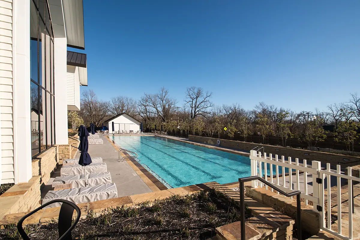 Outdoor pool scene on a sunny day, with clear blue water and surrounded by stone tiles. Umbrella-covered lounge chairs line the poolside, near a white building.
