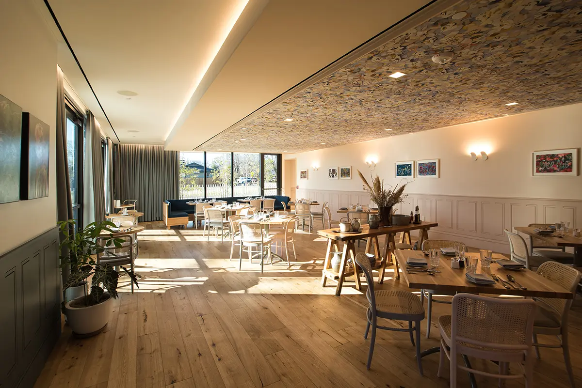 Bright restaurant interior with wooden floors and furniture. Large windows let in natural light. Art adorns walls; plants and decor create a cozy feel.
