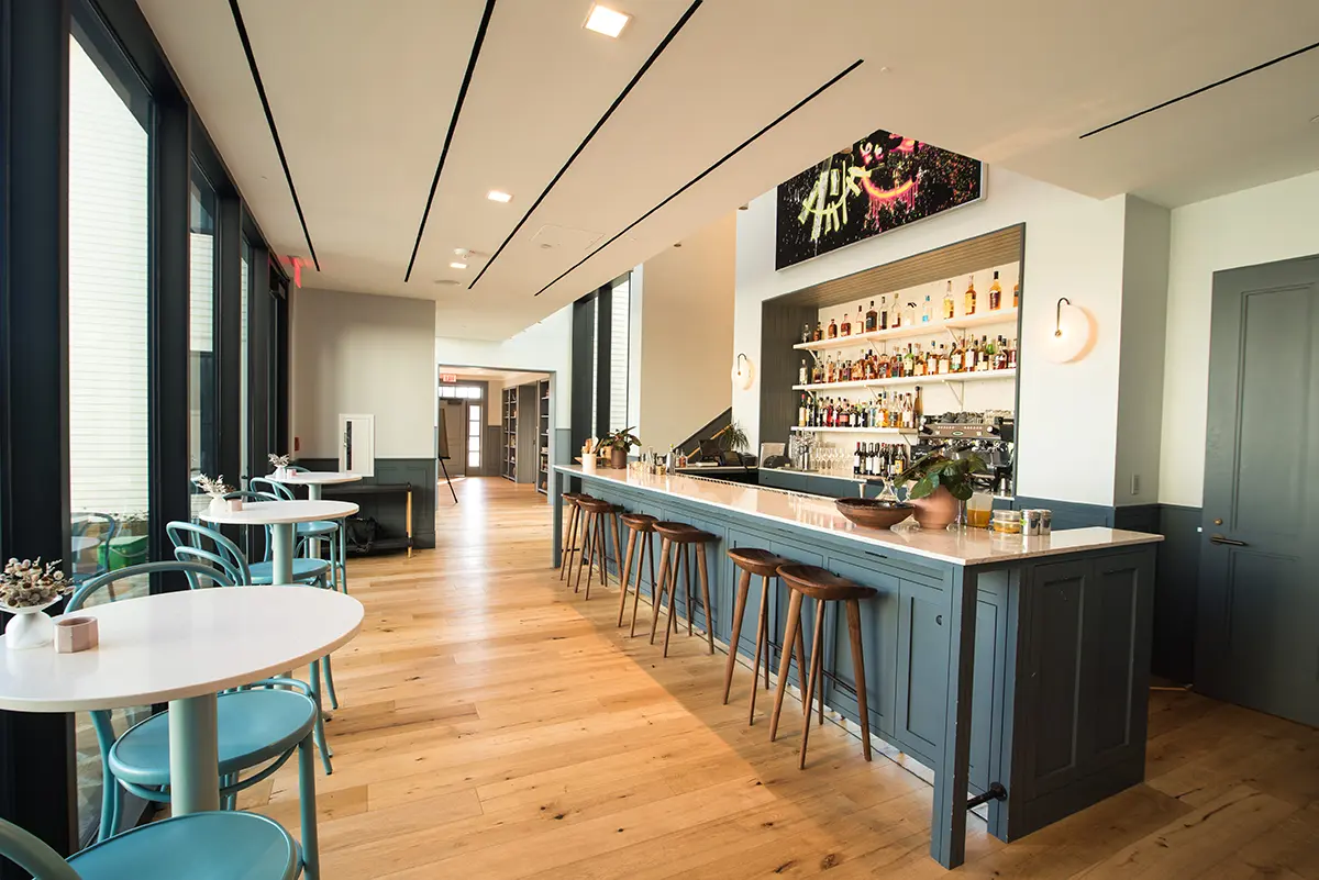 Modern bar interior with a long, blue counter, wooden stools, and shelves of bottles. Natural light streams through large windows. Warm, inviting ambiance.
