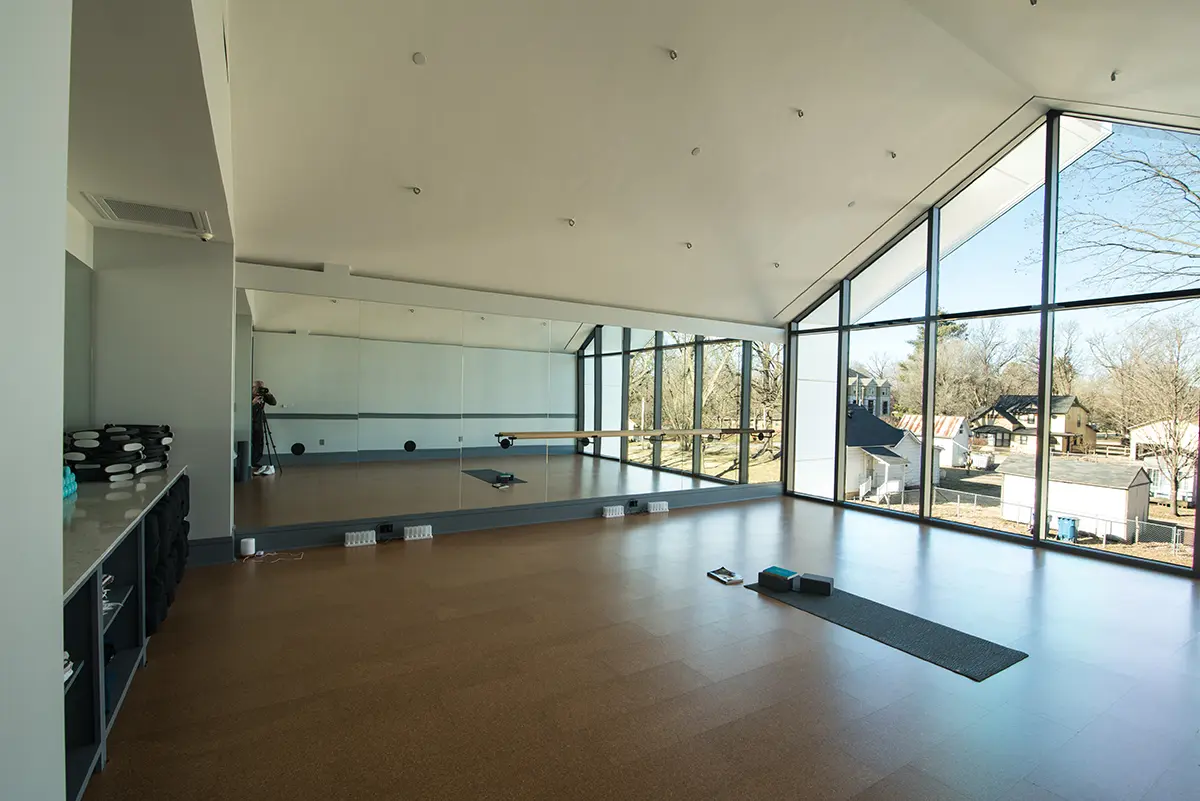 Spacious yoga studio with a wall of mirrors and large windows revealing a sunny outdoor scene. Yoga mat and blocks are on the polished wooden floor.