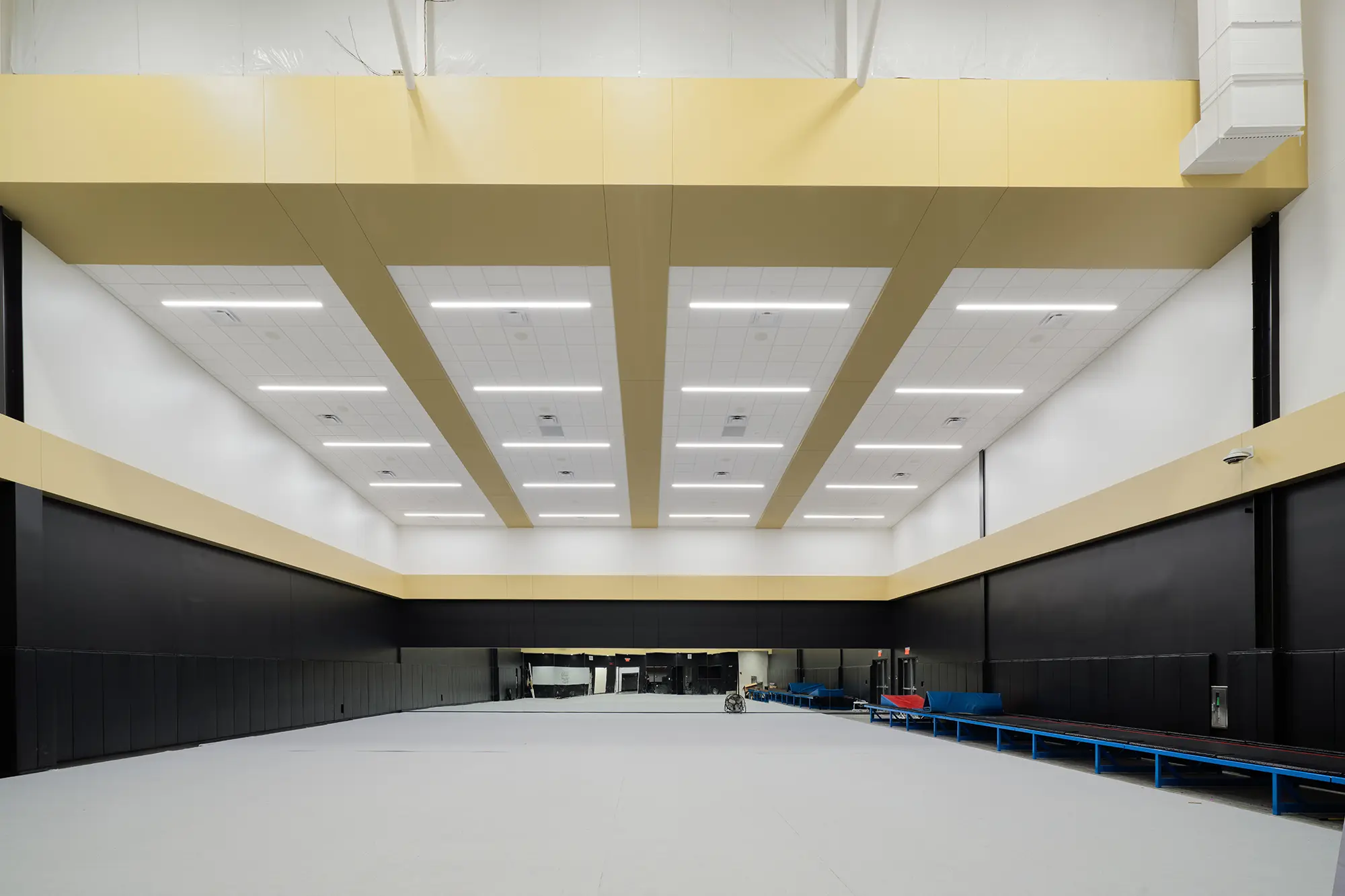 Spacious, empty warehouse with high ceilings and bright overhead lights. Features black walls, a beige band, a large mirror, and blue benches along the right side.