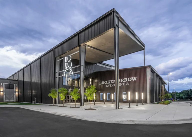 Modern event center with sleek black facade, large glass entrance, and illuminated "Broken Arrow Event Center" signage. Evening sky adds a dramatic backdrop.