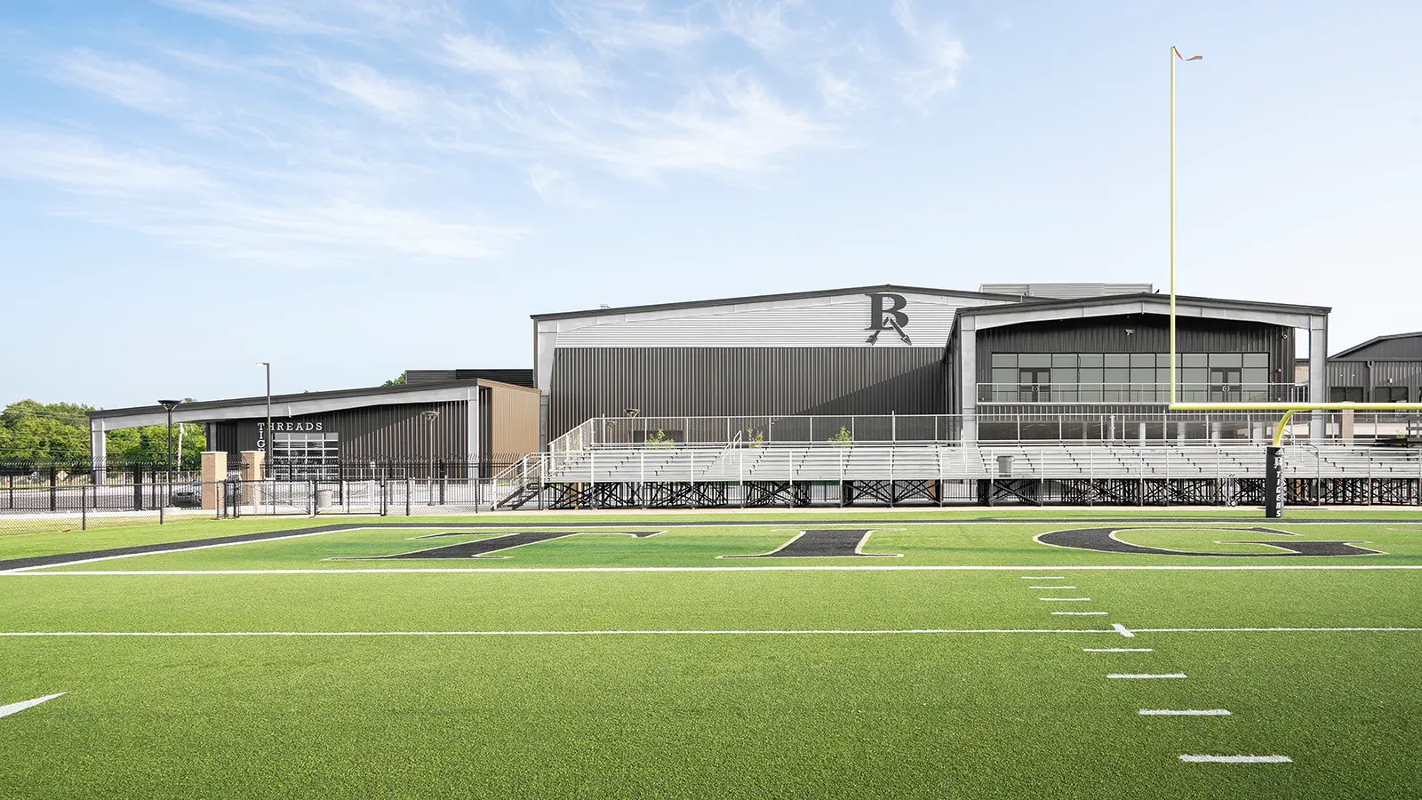 A modern sports complex with a green turf football field in the foreground, bleachers, and a large building in the background under a clear blue sky.
