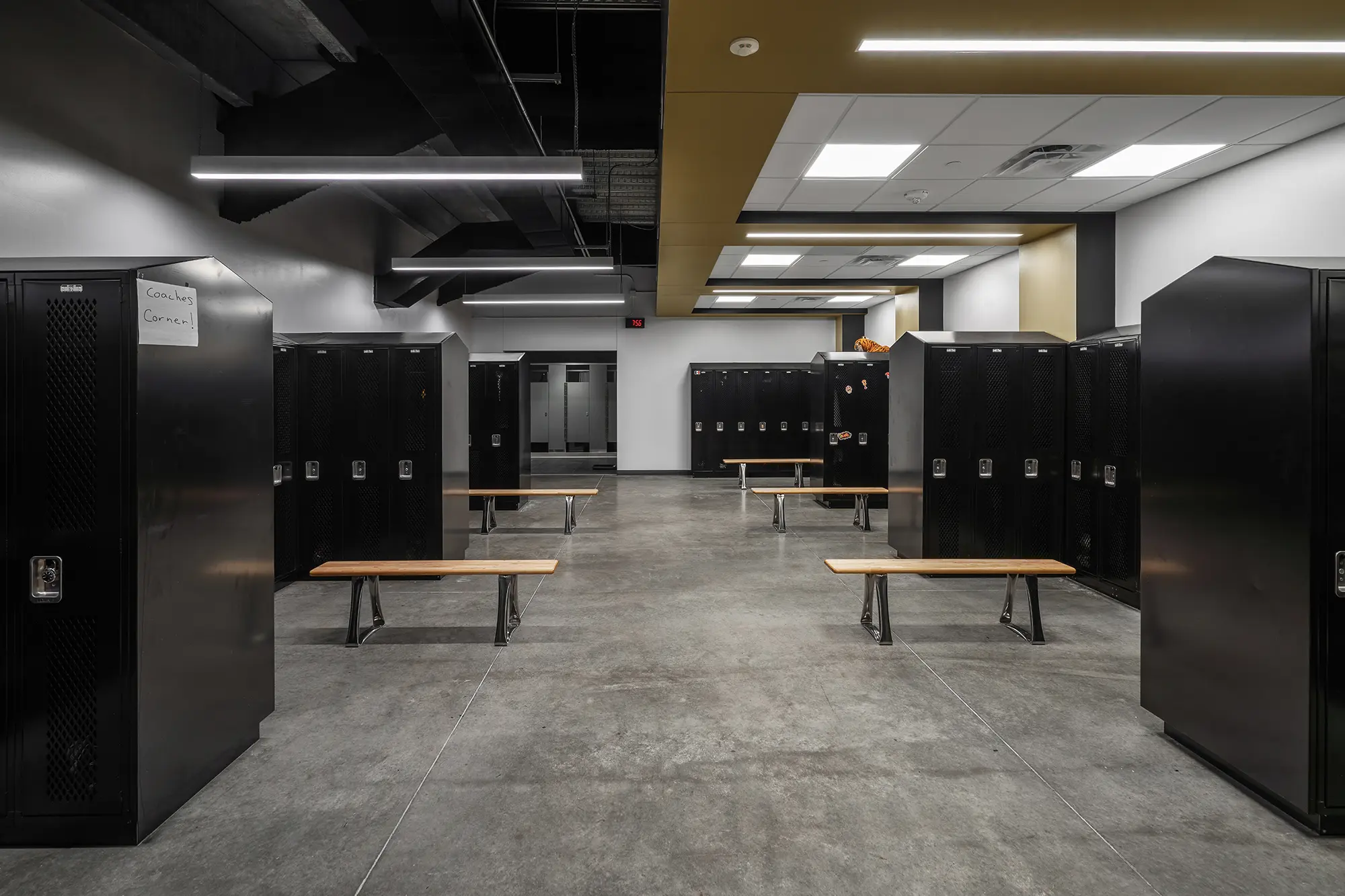 Spacious locker room with black metal lockers and wooden benches on a gray concrete floor. Ceiling lights create a clean, organized atmosphere.