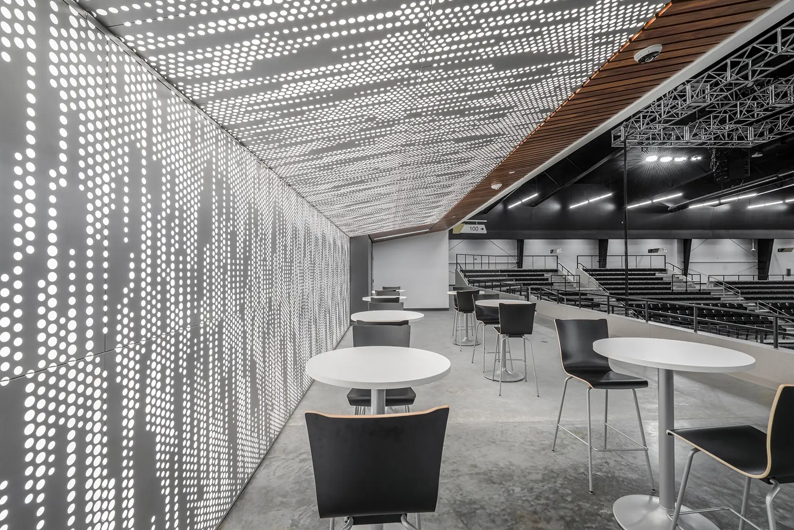 Modern interior with white circular tables and black chairs on a concrete floor. Perforated walls and ceiling create a geometric light pattern.