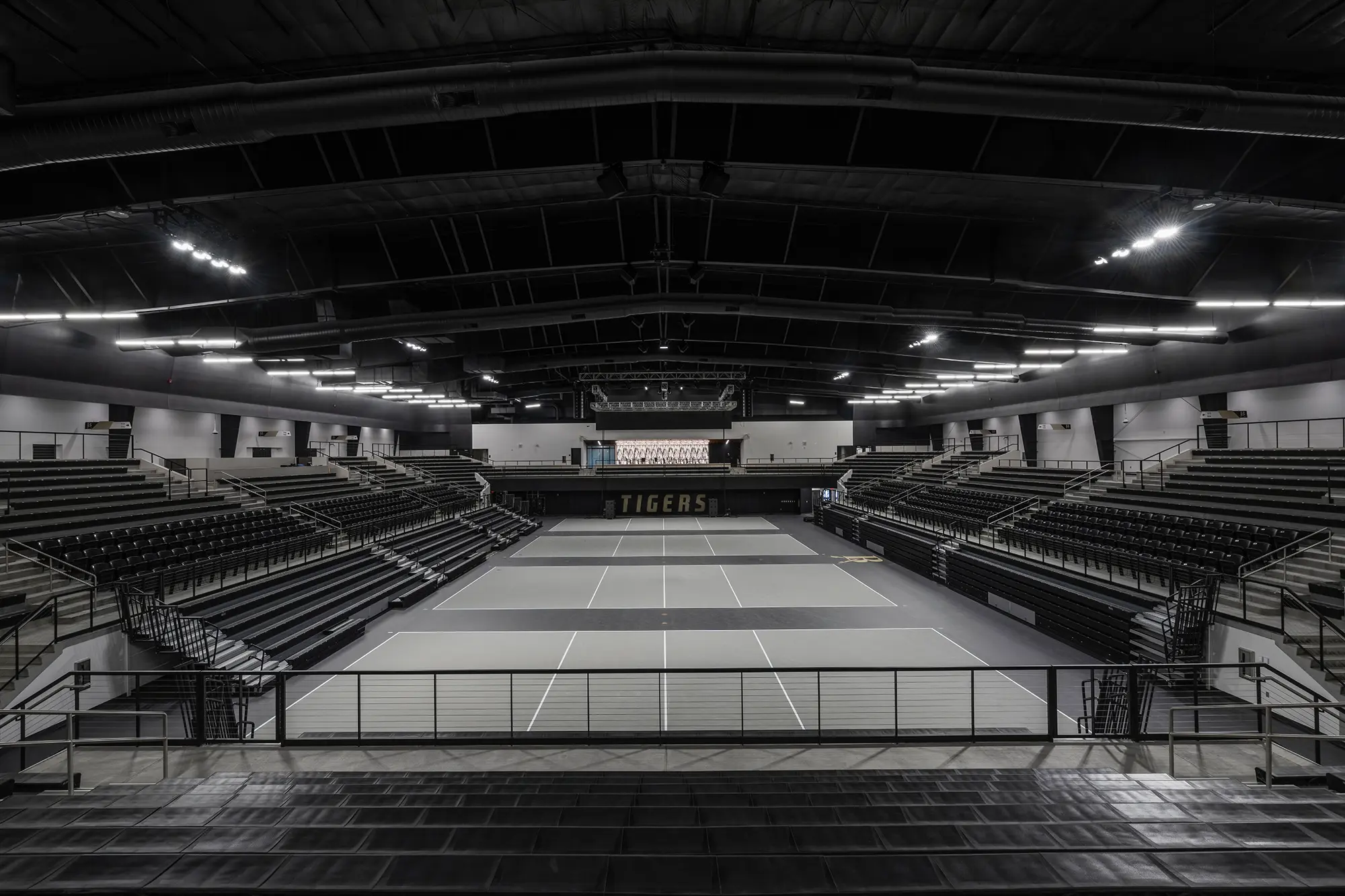 Wide view of an empty indoor sports arena with multiple tennis courts. Dim lighting creates a calm atmosphere. "Tigers" signage visible on the far wall.