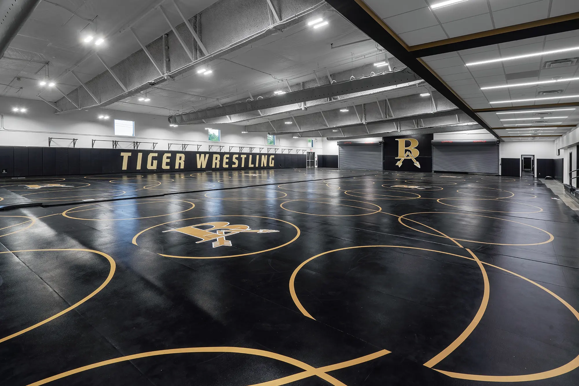 Large wrestling gymnasium with multiple black mats featuring yellow circles and "BR" logos. Walls show "Tiger Wrestling" in bold letters. Bright lighting.