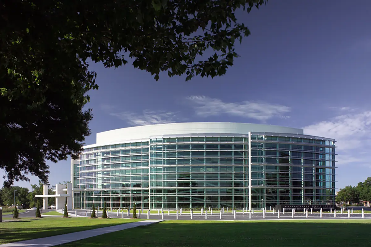 Modern glass office building with a curved facade under a clear blue sky. Surrounded by green grass and trees, conveying a sense of openness and innovation.