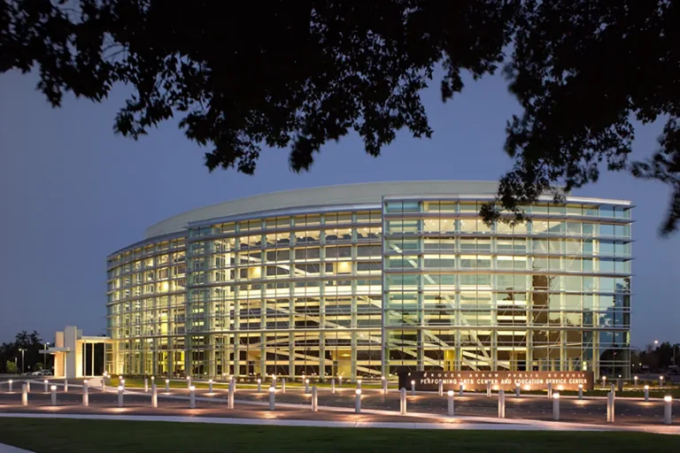 Modern, glass-walled building with curved architecture lit from within at dusk. Surrounded by trees, emitting a calm, inviting ambiance.