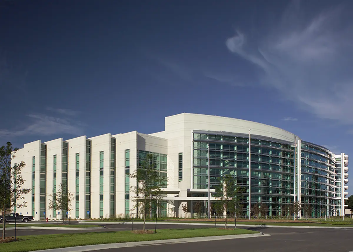 A modern building with a curved design featuring glass and white panels stands against a clear blue sky. A few small trees are planted in front.