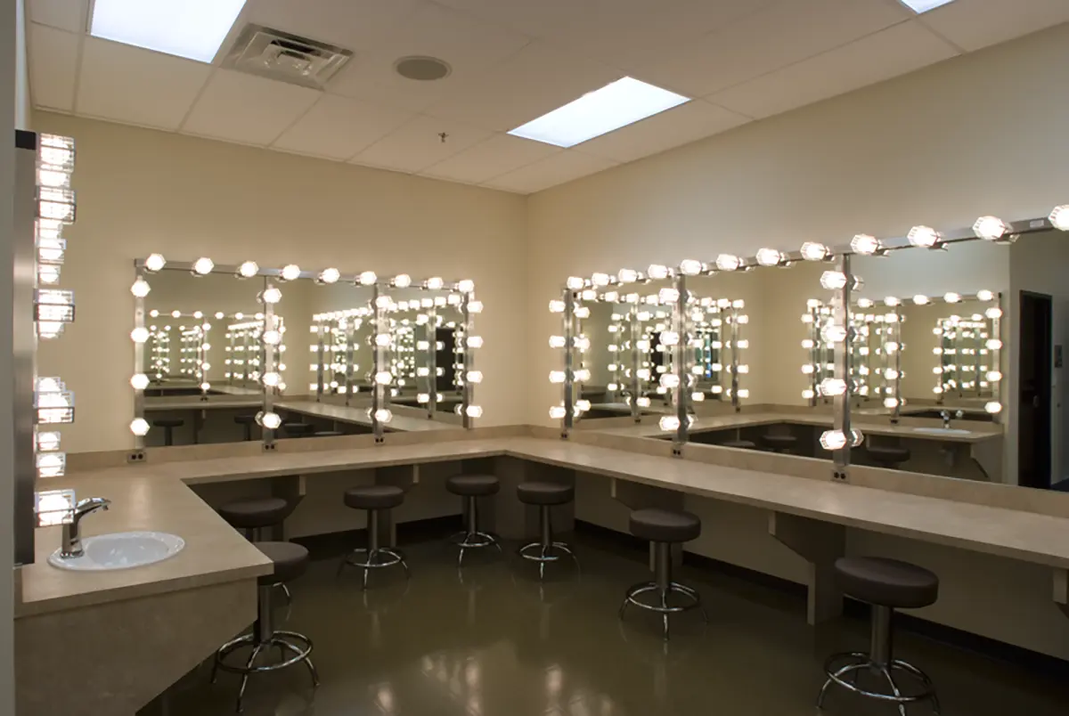 Brightly lit dressing room with large mirrors framed by bulbs, beige countertops, round stools, and a sink. Reflects a calm, professional ambiance.