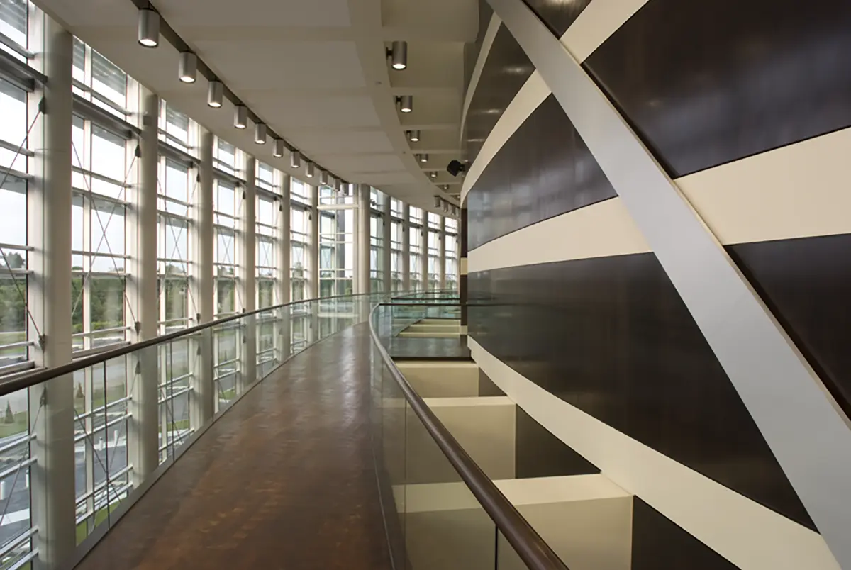 A modern interior features a curved, glass-railed walkway and large windows on the left, with a dark, striped wall on the right. The atmosphere is sleek and spacious.