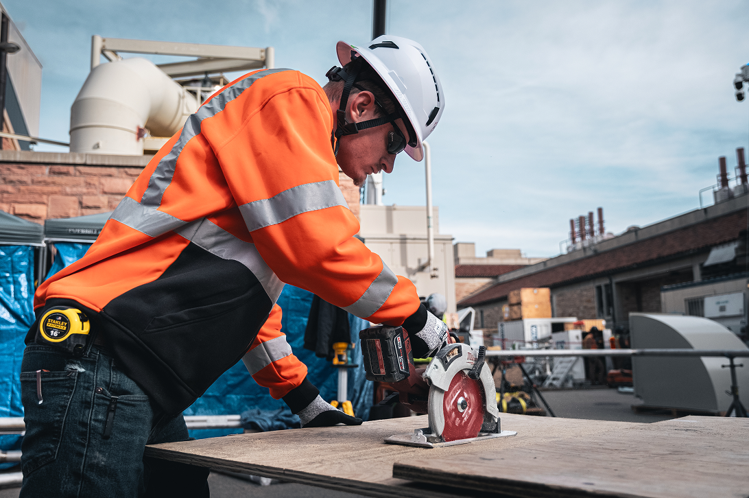 A construction worker in a bright orange safety jacket and helmet uses a circular saw on a wood plank at an industrial site, conveying focus and precision.
