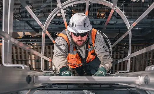 Construction worker wearing a hard hat and orange vest climbs a metal ladder with focus and determination, framed by an industrial structure.