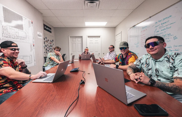 Six people in colorful shirts and sunglasses sit around a conference table with laptops. The mood is relaxed and playful. A whiteboard is visible.