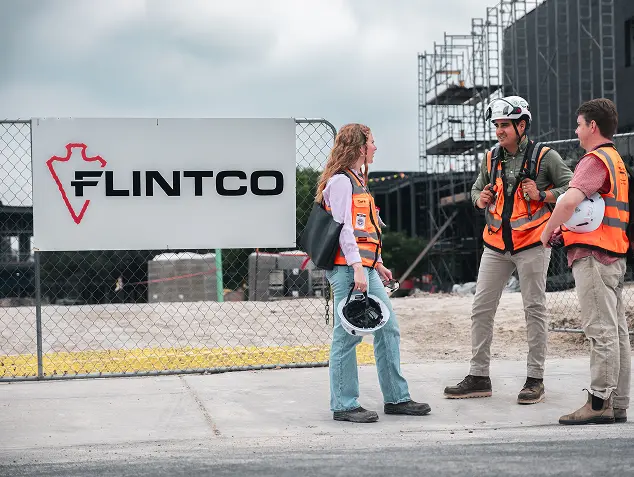 Three construction workers in orange vests converse at a building site. One holds a helmet. A sign reads "FLINTCO." The mood is professional and collaborative.