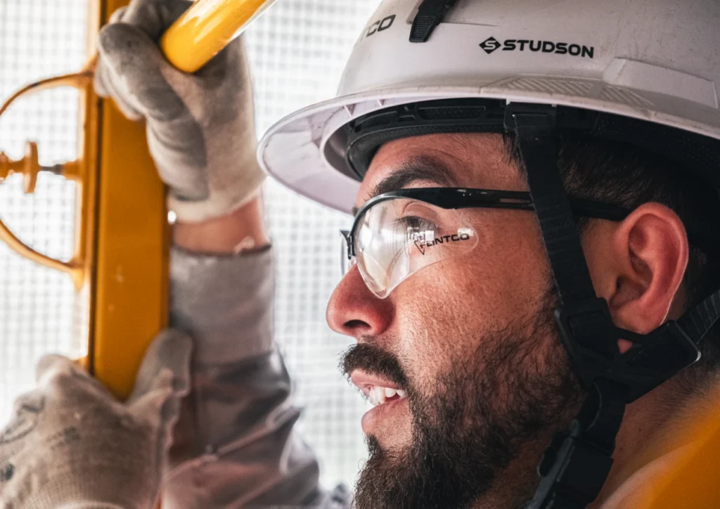 Close-up of a construction worker wearing protective glasses and a hard hat, gripping a yellow handrail. He appears focused and determined.